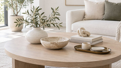 Styled coffee table with vases, bowls, trays, books, and decorative accessories in a bright neutral setting